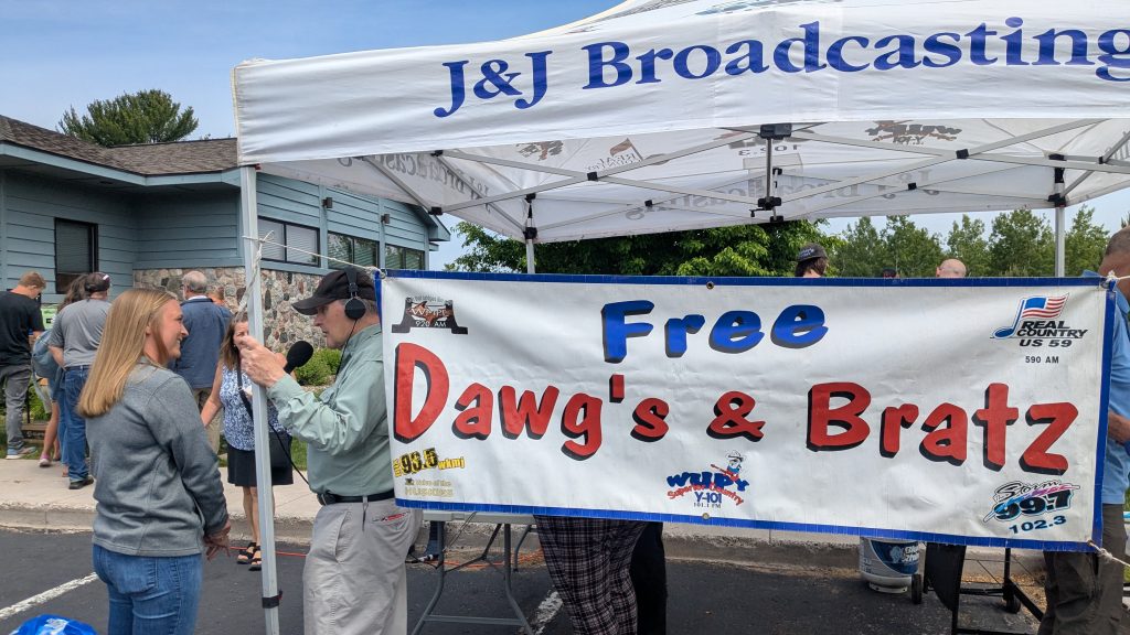 Lender, Bailey, is interviewed by Mitch Lake on live radio broadcast. Large "Free Dawgs & Brats" banner hangs from tent.